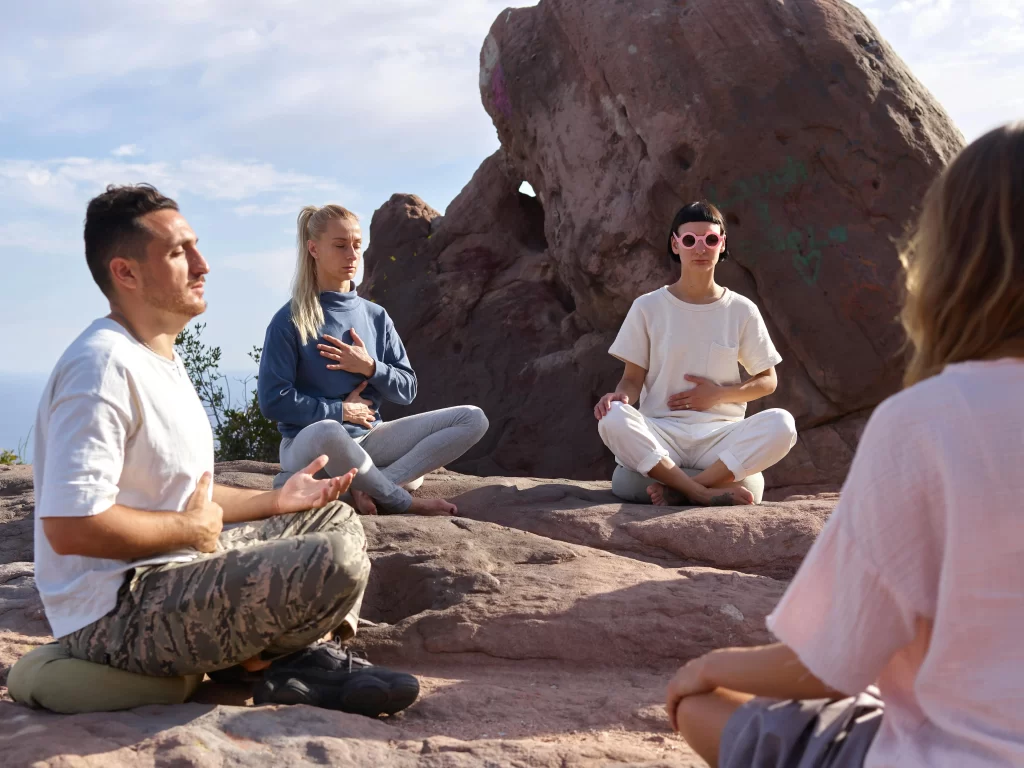 Egy néhány főből álló csoport napos időben a szabadban, egy sziklás részen, körben ülnek és mindfulness légzésmeditációt gyakorolnak. 
