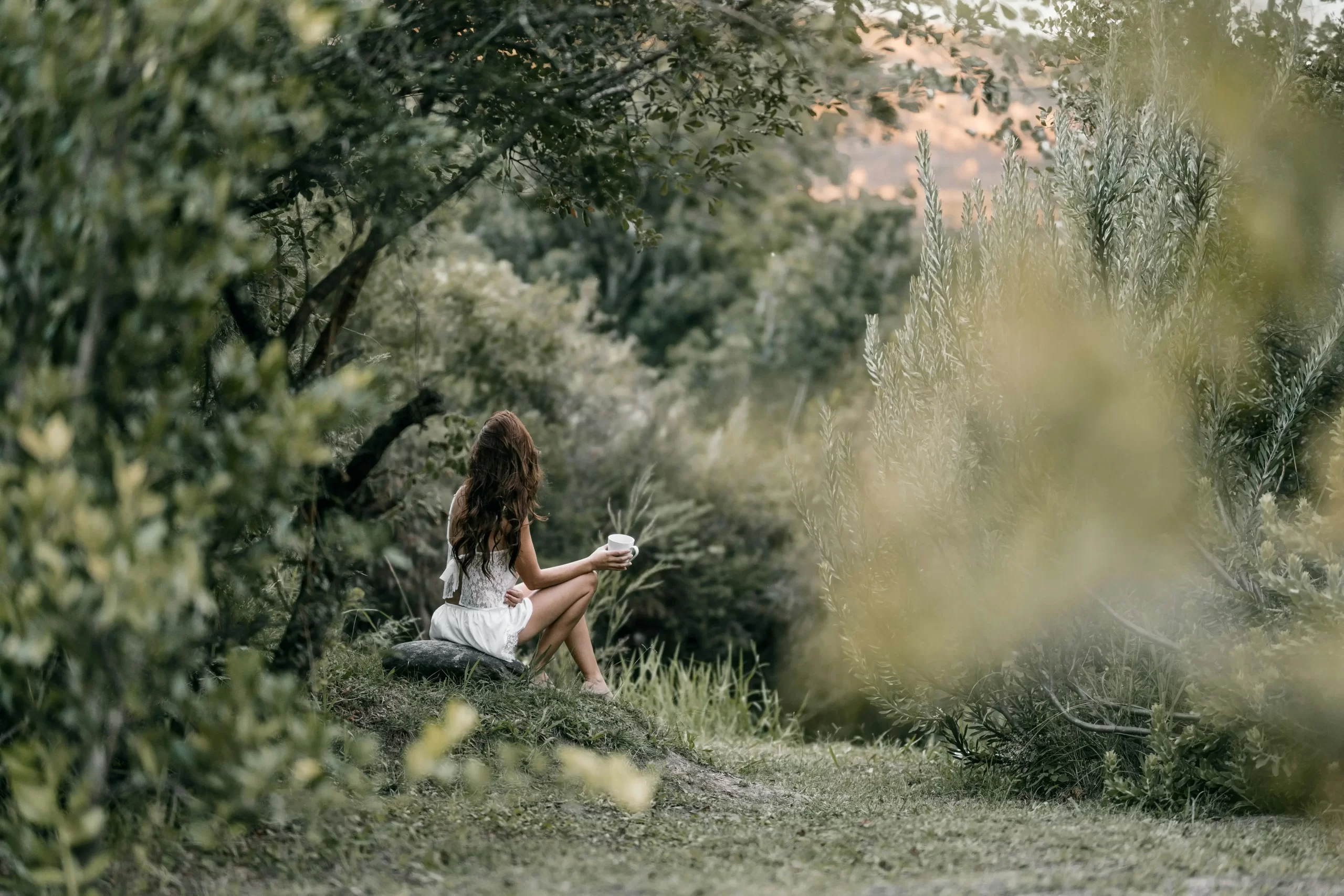 Egy nő a természetben, a kamerának háttal ülve, feher kötött ruhában mindfulness módon kávézik.