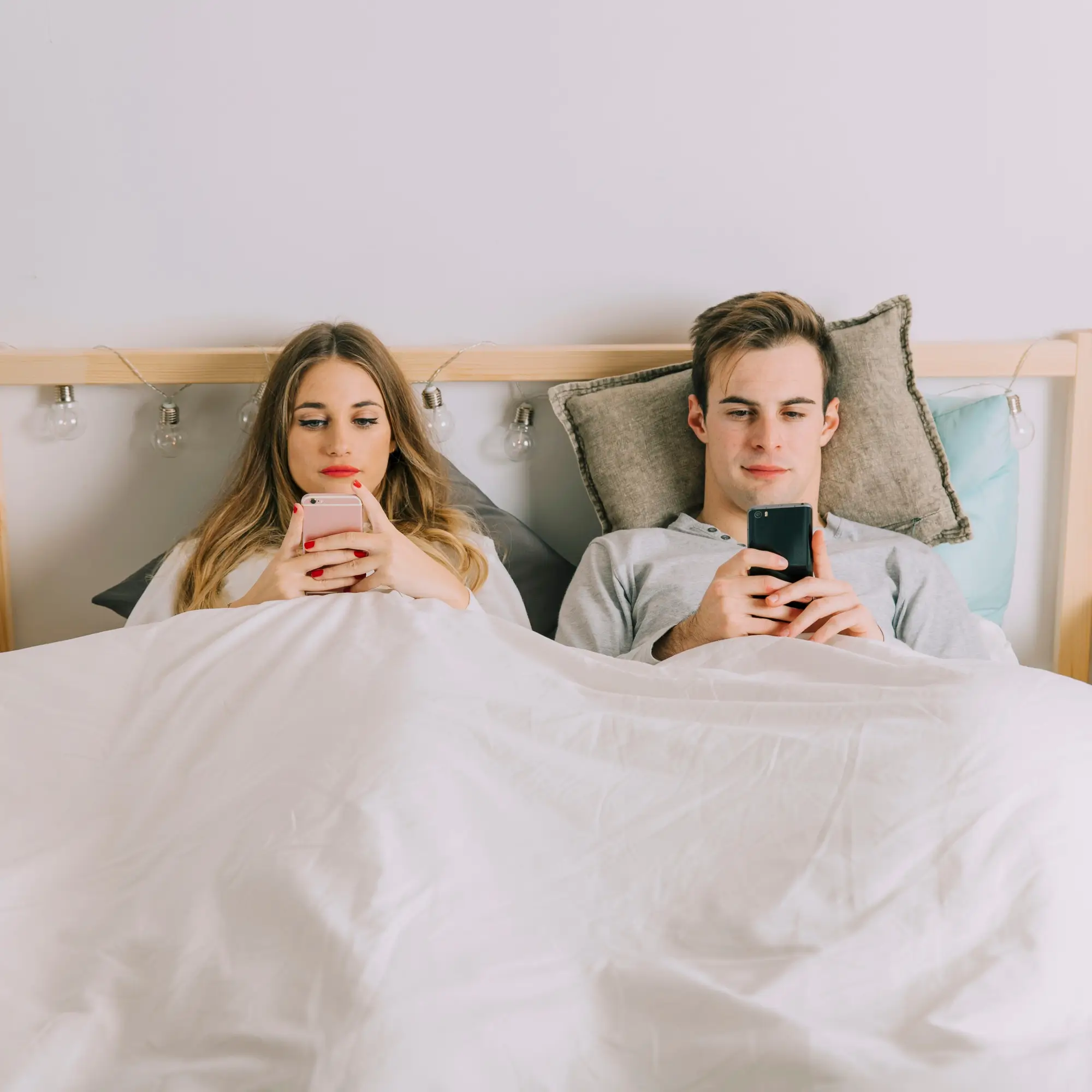 Egy férfi és egy nő fekszenek az ágyban, mindkettő kezében okostelefon, talán mások profilját nézegetik, vagy képeit lájkolják, ami az online hűtlenség jele is lehet.